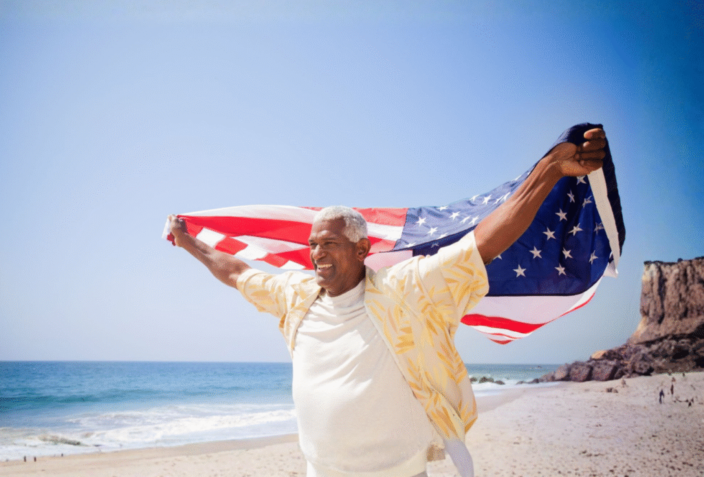 Smiling older man holding an American flag behind him on a sunny beach.